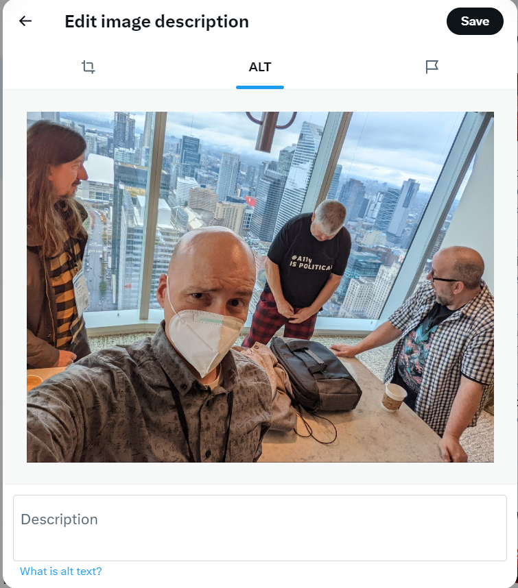 Screenshot of the 'Edit image description' interface on X, a social media platform. The interface displays a photo taken indoors at a high vantage point, showing a group of four people. Adrian in the foreground is taking a selfie while wearing a face mask. Behind him, two people, Jonny and Patrick are looking at another person Steve, who is fiddling with his trousers. The background shows a cityscape with tall buildings. Below the image is a text box labeled 'Description' for adding alt text, with a 'Save' button in the top right corner.